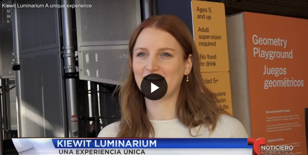 Major Gifts Officer, Mariana Nieto is being interviewed at Kiewit Luminarium, smiling while speaking. The news banner at the bottom says "KIEWIT LUMINARIUM / UNA EXPERIENCIA ÚNICA," with the "Noticiero Nebraska" logo in the corner.